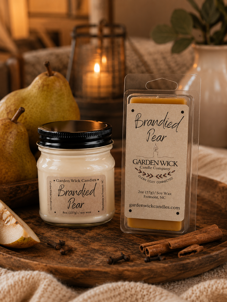 A jar candle and a wax melt package labeled Brandied Pear by Garden Wick Candle Company, styled on a wooden surface with pears, cinnamon sticks, cloves, and a warm lantern in the background.